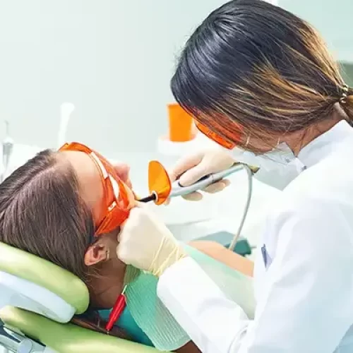A dentist applying a dental sealant to a young patient's teeth using a curing light while the patient wears protective glasses in a dental clinic.